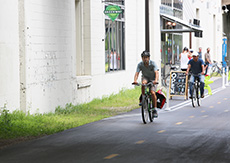 Two bicyclists ride on a paved trail in front of an industrial building. A Midtown Greenway sign hangs above them.