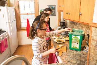 Girls and mom in kitchen putting food scraps into organics recycling container