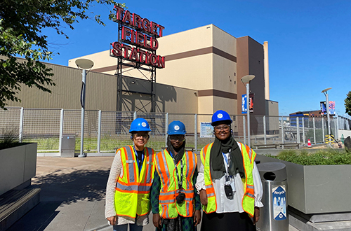 Three green pathways interns in hard hats and safety vests standing in front of Hennepin Energy Recover Center and Target Field sign