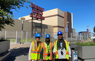 Three green pathways interns in hard hats and safety vests standing in front of Hennepin Energy Recover Center and Target Field sign