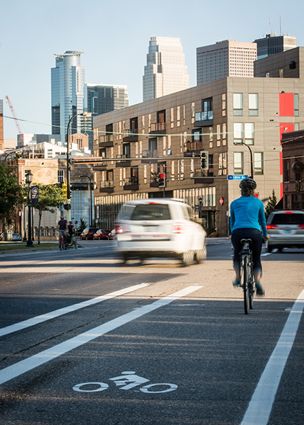 Person biking in bike lane through a neighborhood with a lot of pavement and buildings and few trees or green space