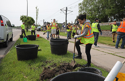 Girl with shovel digging a hole next to a sidewalk during a volunteer tree planting event