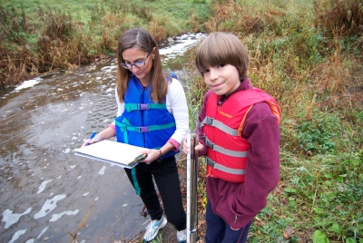 Woman and young boy wearing life jackets standing on edge of stream