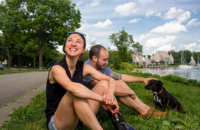 Couple who presents as a man and a woman sitting in the grass with a small black dog next to Lake Harriet with bandshell in the background