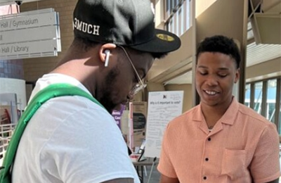 Two people stand indoors; one wears a black cap and uses a tablet, while the other in an orange shirt watches. Signs and posters are visible in the background.