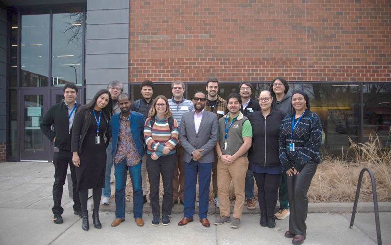 A group of 14 people posed in two rows stands in front of a red brick building.