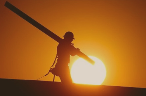 Construction worker working in the heat, carrying a beam with the sun blazing behind them