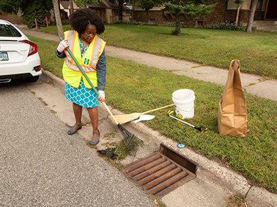 Woman in safety vest with rake cleaning grass clippings out of storm drain