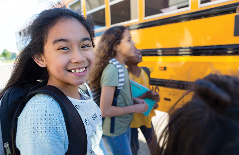 Young girl smiles while a group of students wearing backpacks stand in front of a school bus.