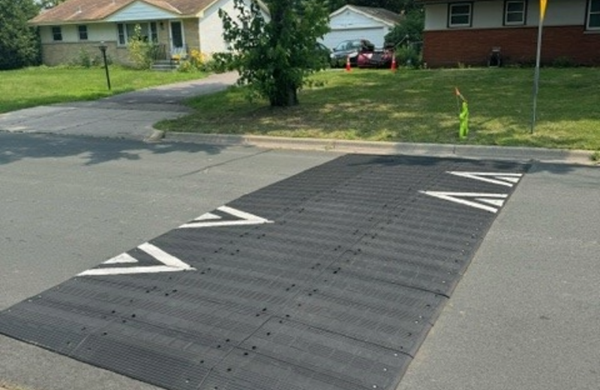 A large black speed bump with white arrow markings spans across a suburban street. A caution flag and parked van are visible, with grass and trees in the background.